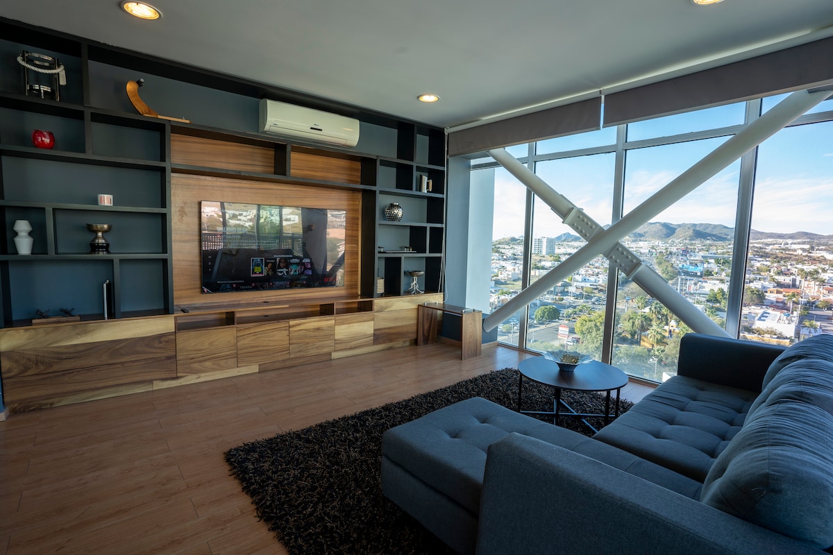 A modern living area is shown, featuring a comfortable gray sofa facing a wall-mounted television. A wooden media console and shelving add functionality, while large windows allow natural light and provide extensive views of the surroundings.