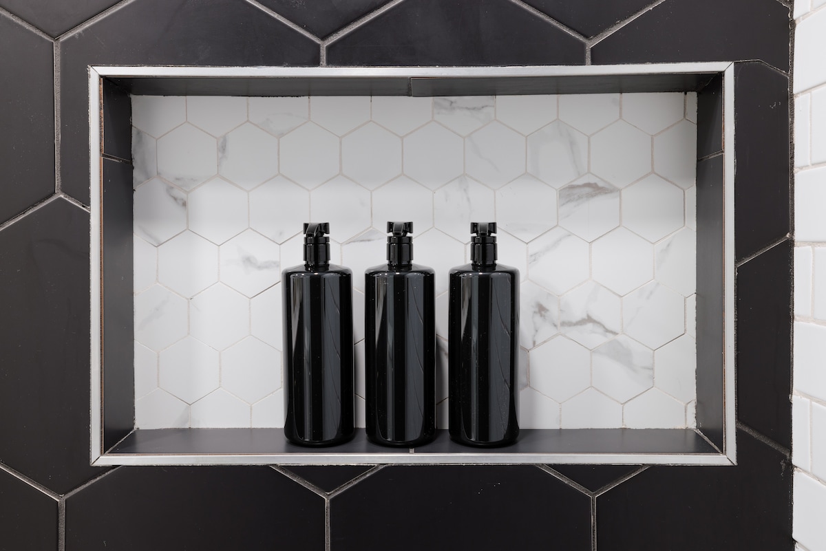 A shower niche displays three sleek black bottles arranged symmetrically against a backdrop of hexagonal tiles. A contrasting section of white tiles with subtle marbling complements the design, creating a modern look.