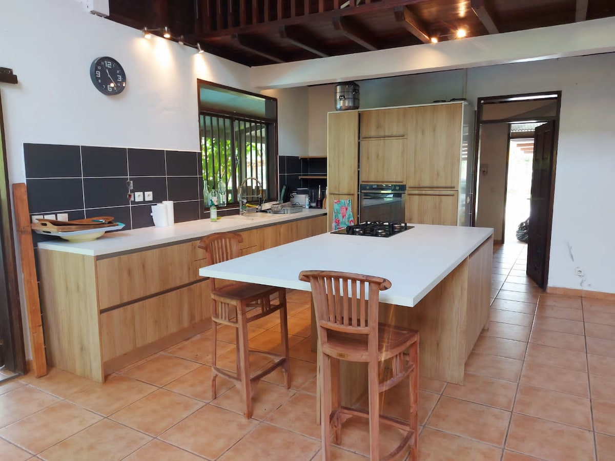 A modern kitchen features a wooden cabinetry and an island with seating for four. The space is well-lit with natural light streaming in through a window, and a clock is mounted on the wall for convenience. An open layout connects the kitchen to adjacent areas.