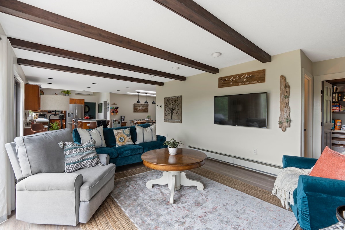 An open living space features comfortable seating arrangements, including a large sectional sofa and accent chairs. A round coffee table sits at the center, surrounded by neutral-toned area rugs. Natural light filters through large windows, illuminating the warm wood beams overhead.