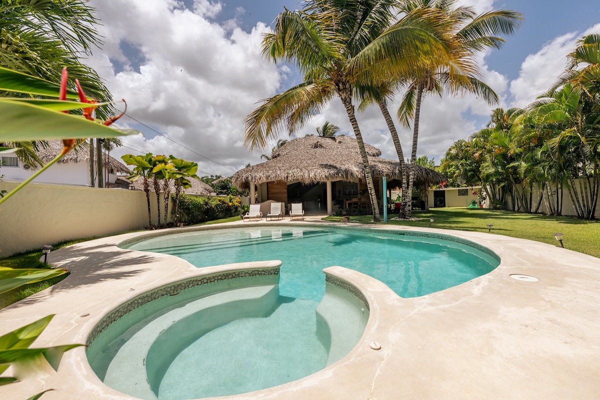 A private swimming pool is set against a backdrop of palm trees and a thatched-roof gazebo. The pool features a spacious area and a built-in jacuzzi, surrounded by a well-maintained garden to offer a relaxing outdoor experience.
