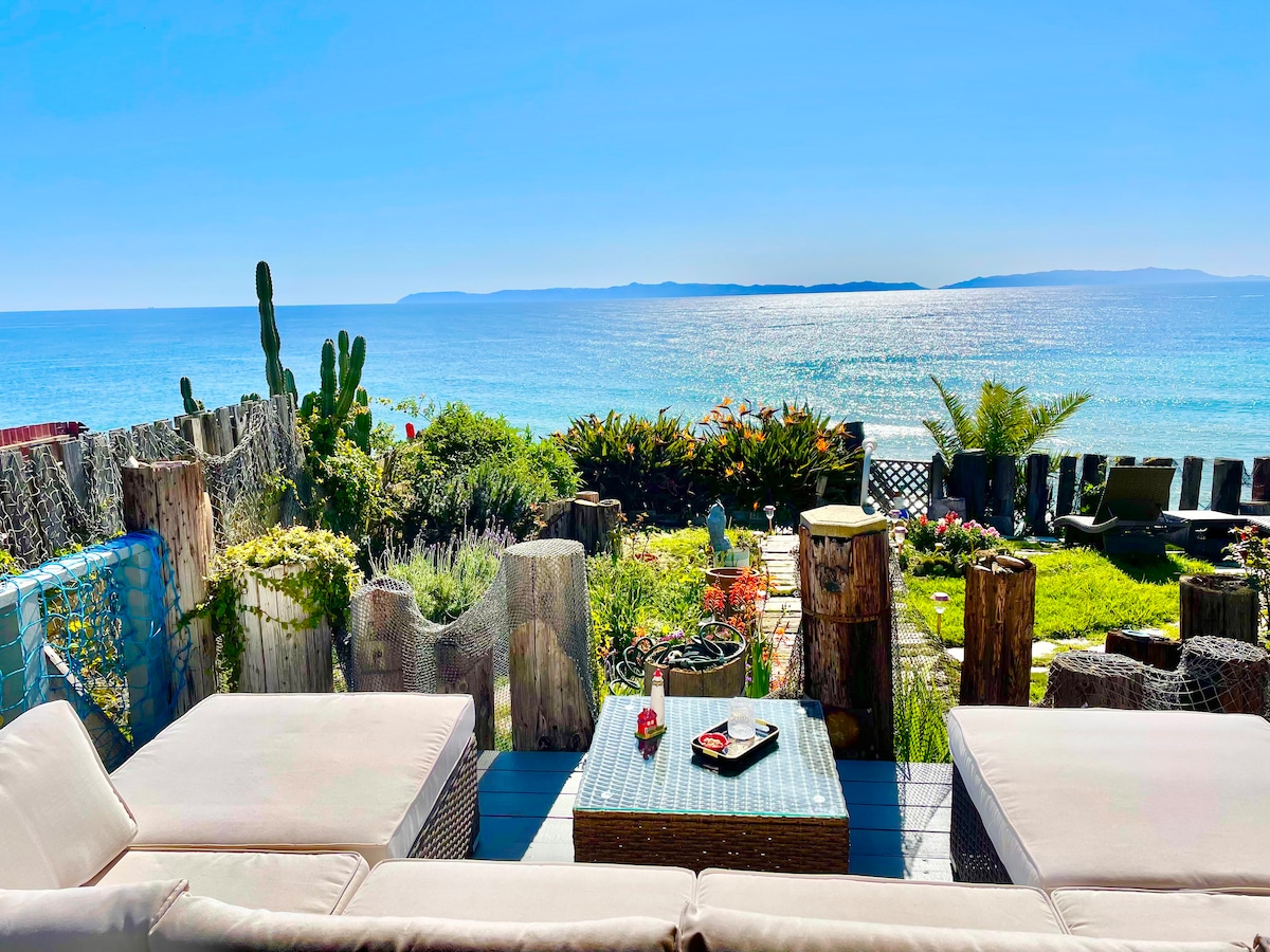 A cozy outdoor seating area is shown with soft cushions on loungers, overlooking vibrant greenery and sparkling blue ocean waters. The view includes distant islands on the horizon. A small table is set with snacks, inviting relaxation and enjoyment of the scenery.