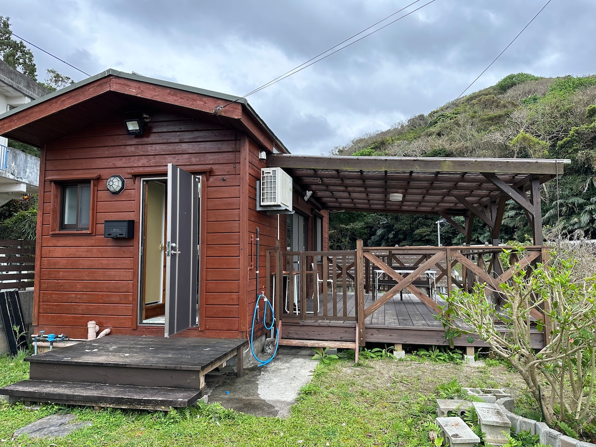 A wooden cabin features a sloped roof and a spacious, covered deck. An air conditioning unit is mounted beside the entrance. The surrounding area includes a small garden with grass and low shrubs. Scenic hills are visible in the background under a cloudy sky.