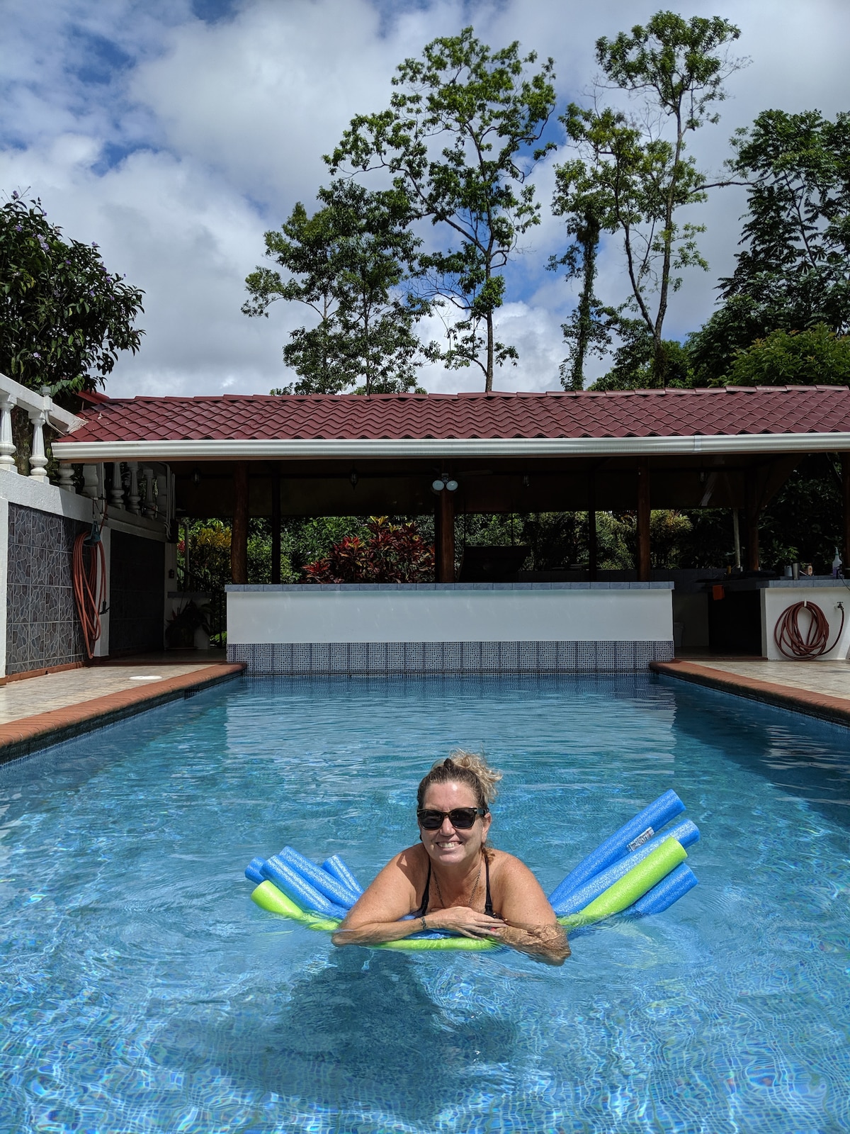 A clear blue swimming pool is surrounded by lush greenery, with tropical trees visible in the background. Colorful pool floats rest on the water's surface, and a covered seating area is positioned at one end of the pool, complemented by vibrant landscaping.