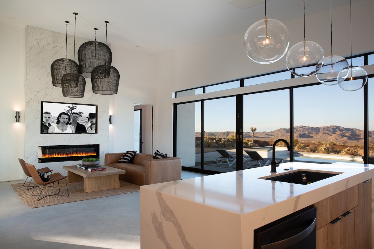 A modern living area showcases a spacious sofa and stylish chairs surrounding a central coffee table on a light rug. Large glass doors offer views of the mountains, while a sleek kitchen island features a contemporary sink. Pendant lights hang above, illuminating the space.