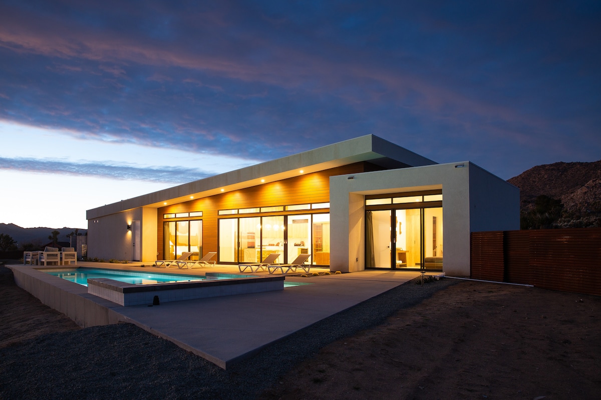 The exterior view of the modern desert retreat showcases large glass windows, allowing natural light to illuminate the spacious interior. The private pool glimmers under the evening sky, complemented by a contemporary lounge area. The landscape features sandy tones, blending with the surrounding mountains.