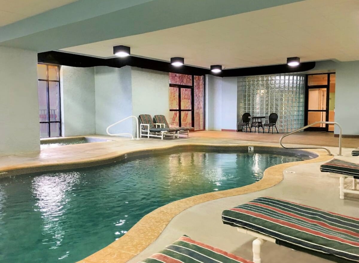 An indoor pool area features a gently curving pool surrounded by comfortable lounge chairs. Natural light filters through large windows, illuminating the space. A small table and chairs are positioned near another wall, offering a cozy spot for relaxation.
