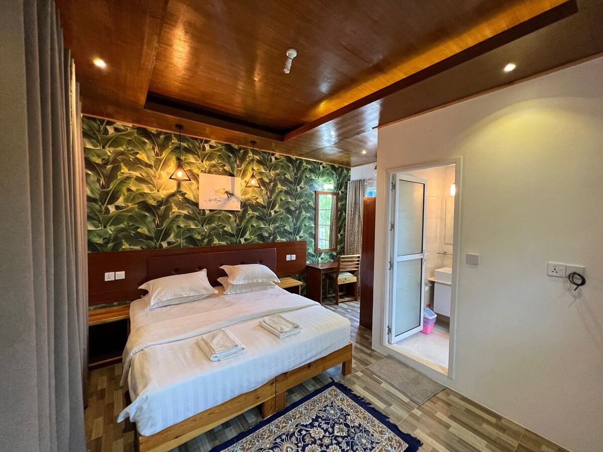 A well-appointed guest room features two spacious beds with neutral linens. A vibrant leafy wallpaper adds character to the room, while a wooden ceiling and flooring enhance the natural feel. An ensuite bathroom is visible through an open door, providing convenient access.