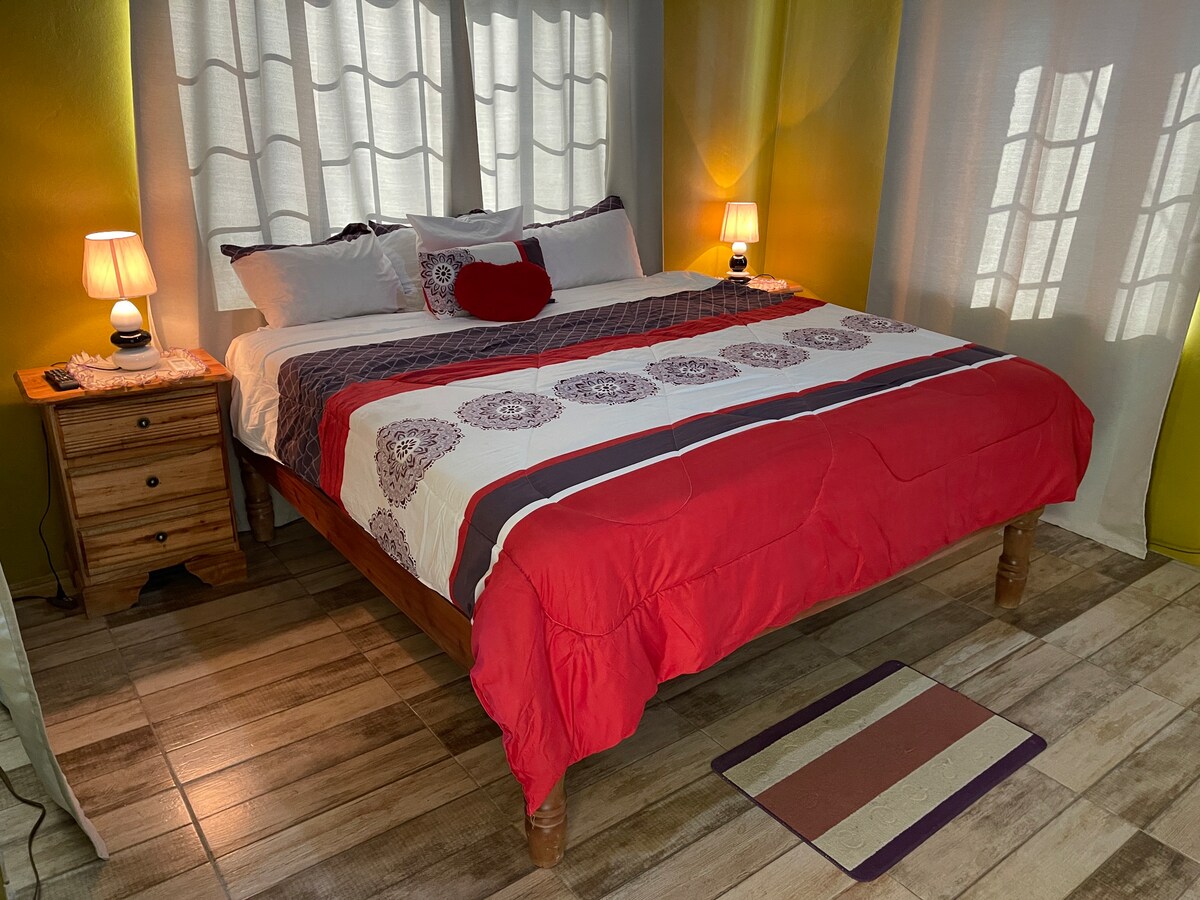 A spacious bed is centered in the room, adorned with a vibrant red and white comforter. Nightstands with lamps flank the bed, providing warm lighting. Sheer curtains allow soft light to filter in, enhancing the inviting atmosphere of the space, with decorative elements along the wooden flooring.