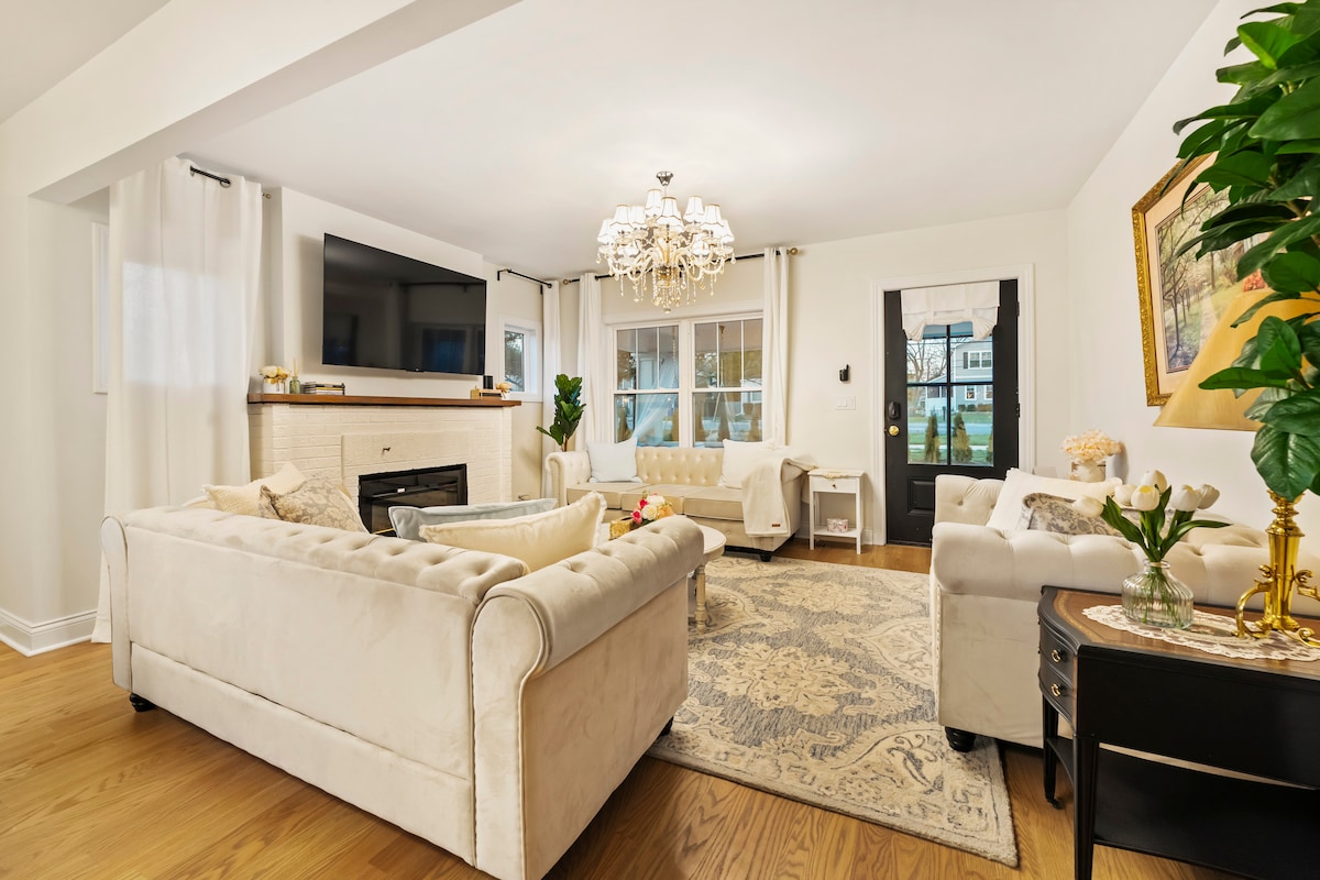 A spacious living area features light-colored sofas arranged around a decorative area rug. A chandelier hangs from the ceiling, providing soft illumination. Large windows bring in natural light, and a cozy fireplace is visible beneath a mounted television. Potted plants add greenery to the space.