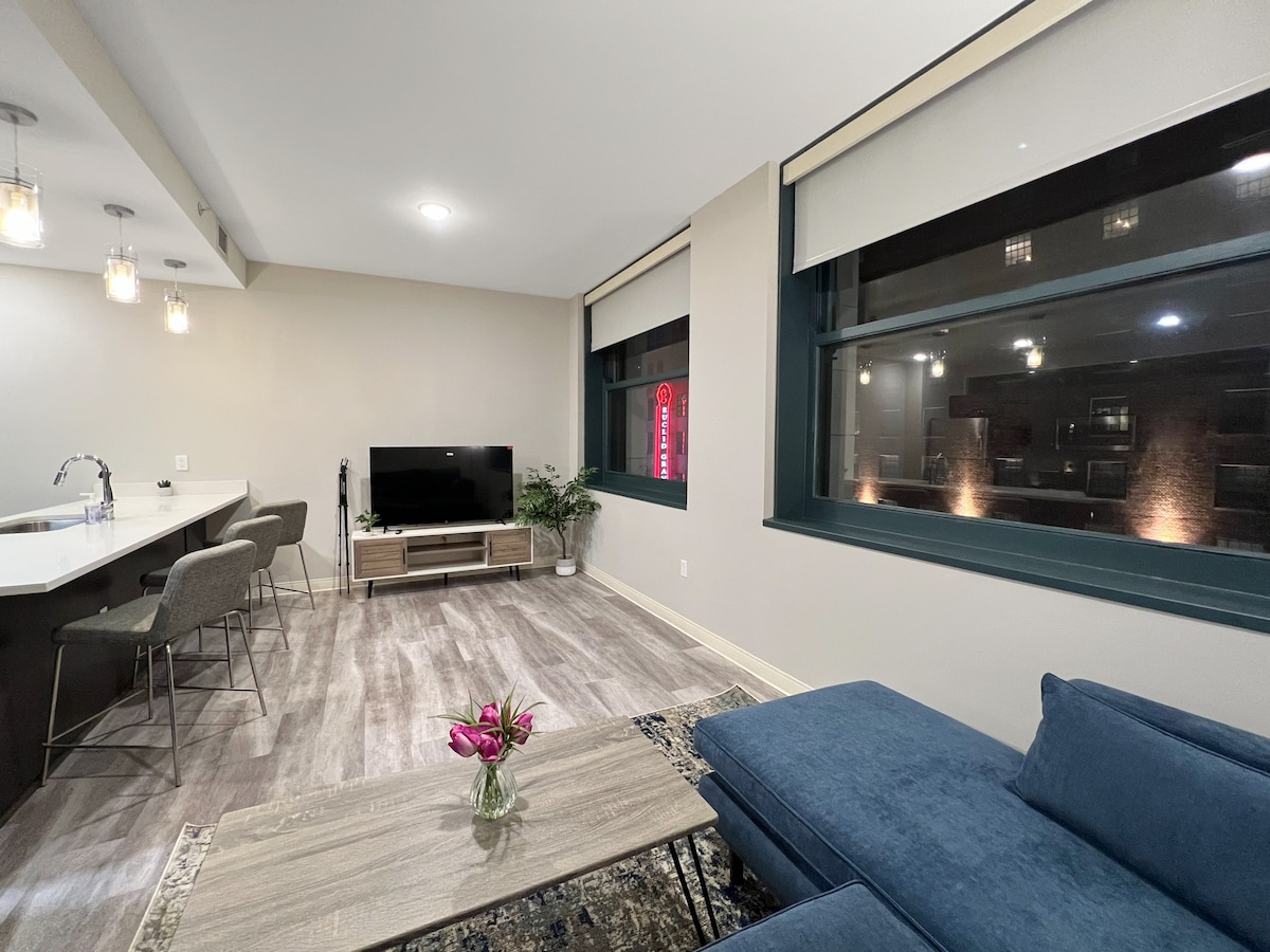 A modern living space features a comfortable sectional sofa positioned near oversized windows, allowing for ample natural light. A sleek TV unit with a flat-screen TV is visible, complemented by a small coffee table adorned with fresh flowers. The open layout includes a dining area with stylish counter stools.