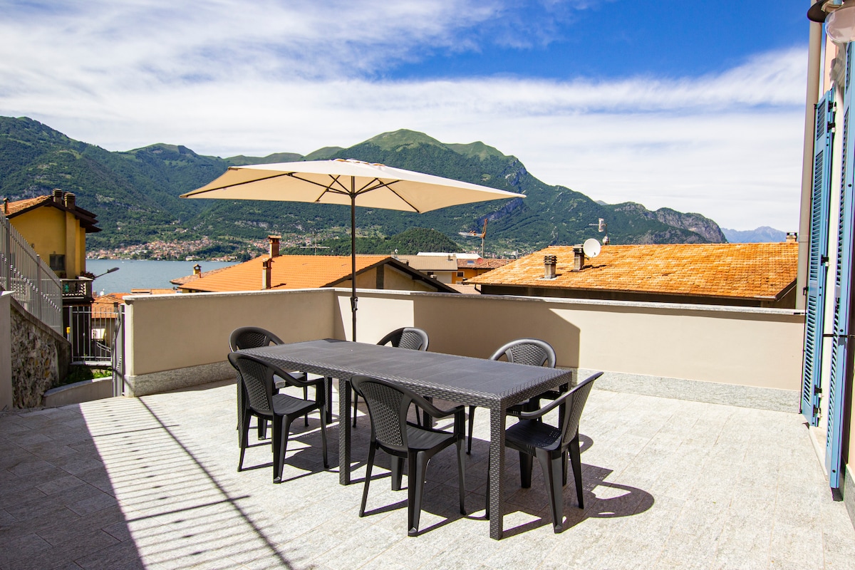 An outdoor terrace is shown, featuring a large dining table surrounded by six black chairs. A large umbrella provides shade, and the scenic view includes mountains and a lake in the background under a partly cloudy sky.
