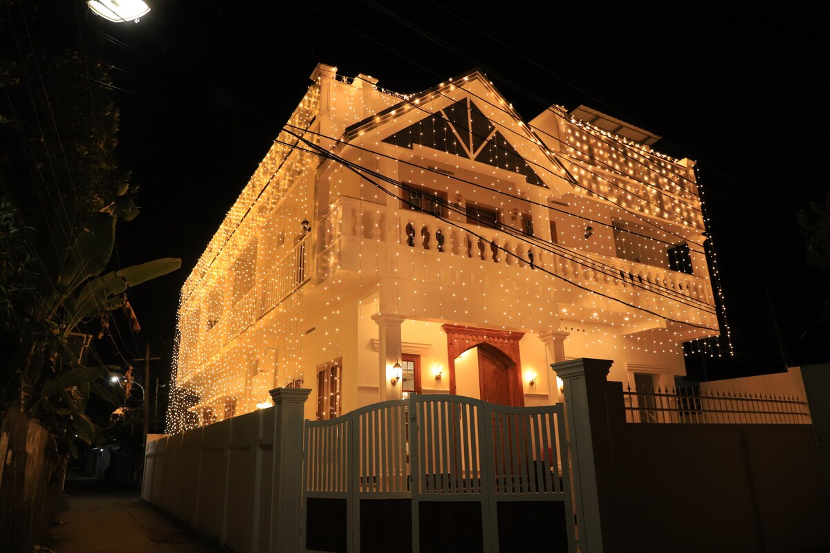 A well-lit building is adorned with numerous lights, creating a festive atmosphere at night. The architecture features a combination of traditional and modern elements, with decorative balconies and a welcoming entrance. A white fence surrounds the property, contributing to the overall charm.