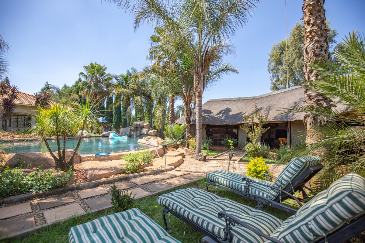 Lounge chairs with striped cushions are arranged in the foreground, facing a tranquil pool surrounded by palm trees and lush greenery. A thatched-roof structure is visible in the background, enhancing the outdoor space's inviting ambiance.