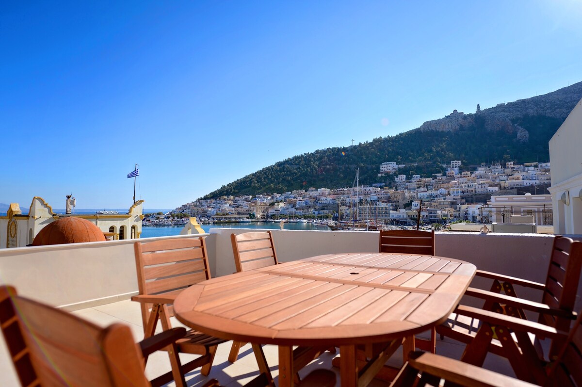 A spacious outdoor terrace is featured, furnished with a large wooden dining table surrounded by several matching chairs. The view extends towards the harbor and hillside, showcasing the charming buildings and greenery of the surrounding area, all under a clear blue sky.