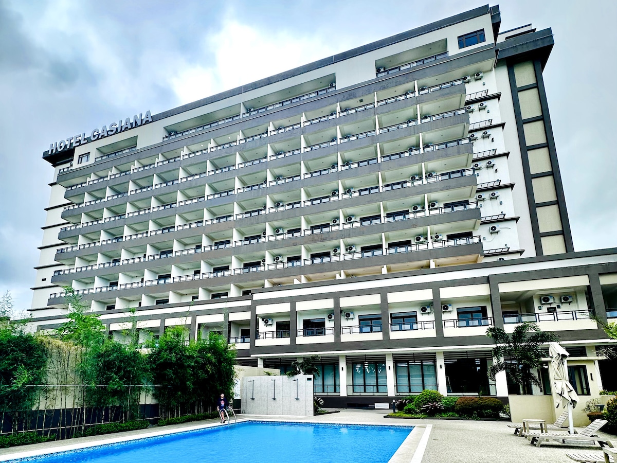 The exterior of Hotel Casiana is depicted, showcasing a modern multi-story building with a sleek facade. A serene swimming pool is located in front, surrounded by landscaped areas. Several air conditioning units are visible along the balconies, enhancing guest comfort.