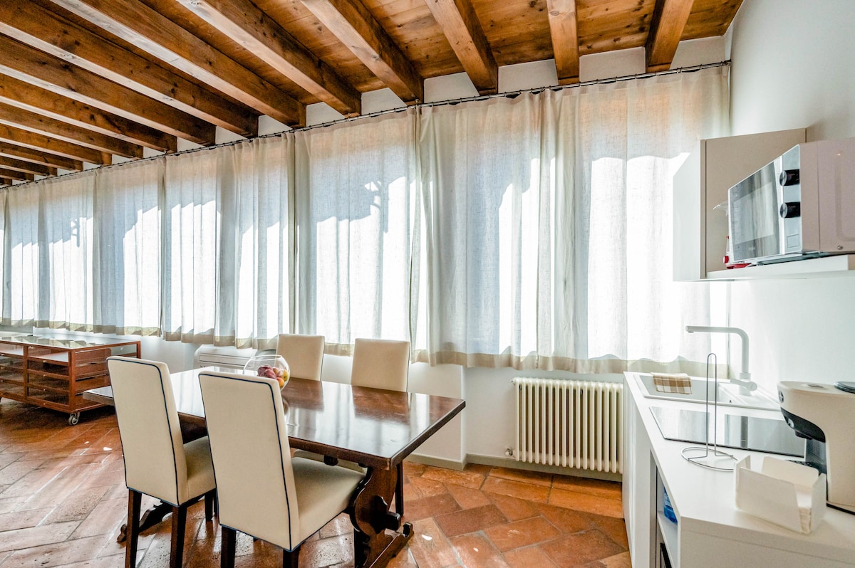 A spacious dining area is illuminated by natural light filtering through floor-to-ceiling windows, adorned with soft, sheer curtains. A large wooden table is surrounded by several light-colored chairs. A modern kitchenette is visible, equipped with essential appliances like a microwave and sink.