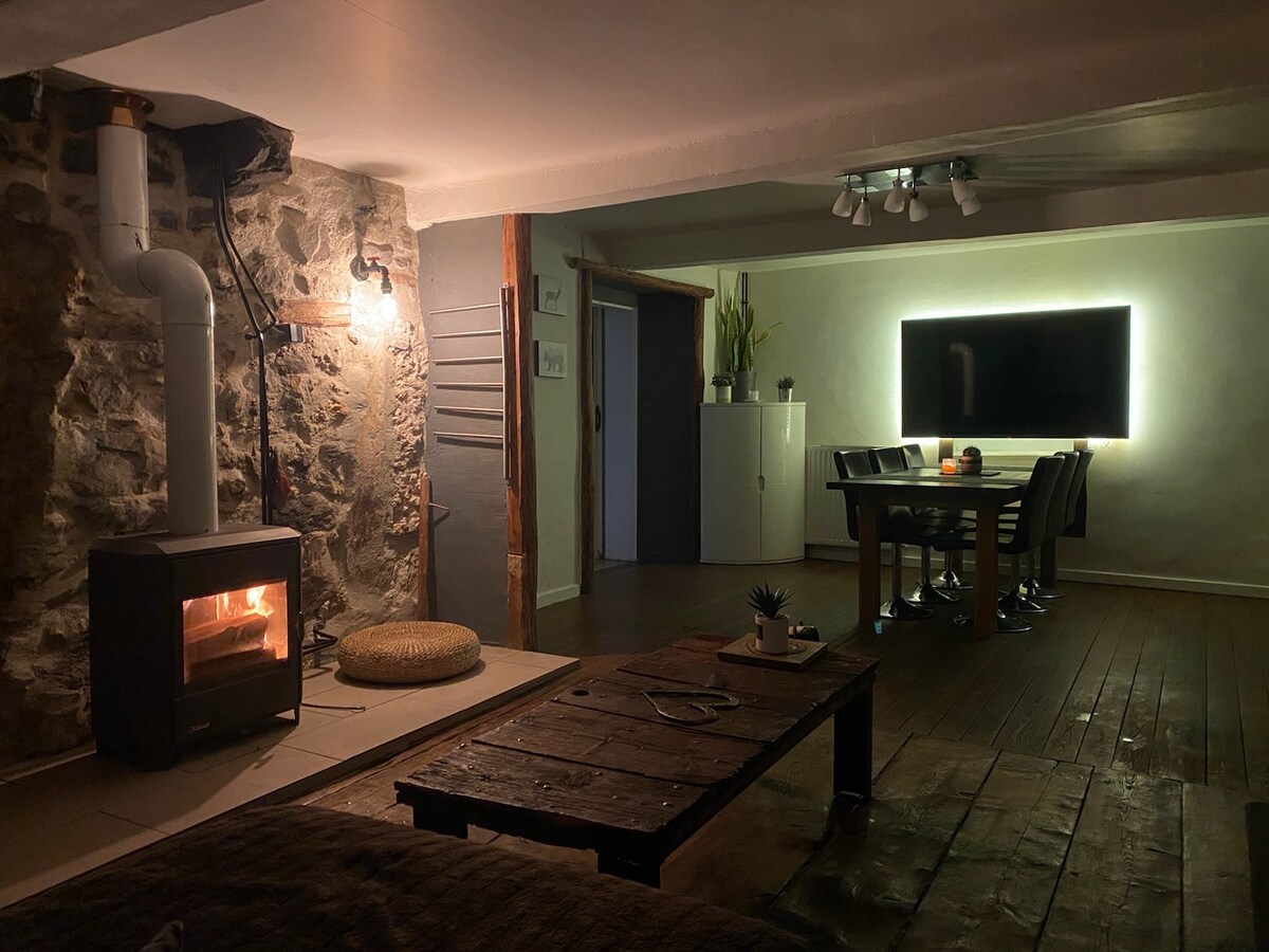 A cozy living space features a wood-burning stove, providing warmth to the room. A rustic wooden coffee table sits near a comfortable seating area. A dining table for six is illuminated by wall-mounted lighting, contrasting with the natural stone walls in a softly lit atmosphere.