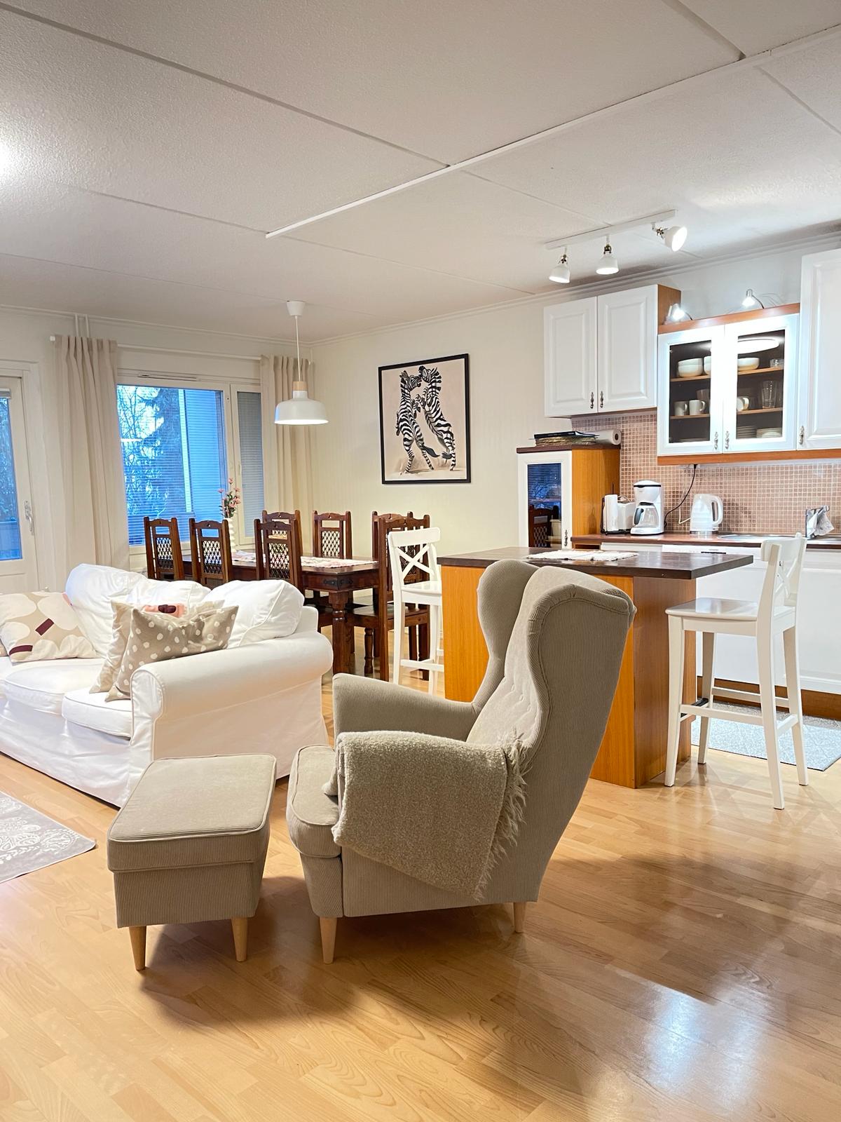 A bright living area displays a comfortable white sofa and a stylish light gray armchair with a footrest. A wooden dining table with six chairs is positioned nearby, and an open kitchen area with white cabinetry is visible in the background.