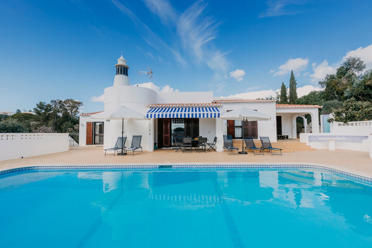 A spacious villa features a clear blue pool in the foreground, surrounded by lounge chairs. The exterior is accentuated by large windows and a striped awning, creating a welcoming outdoor dining area. Lush greenery and trees are visible in the background.