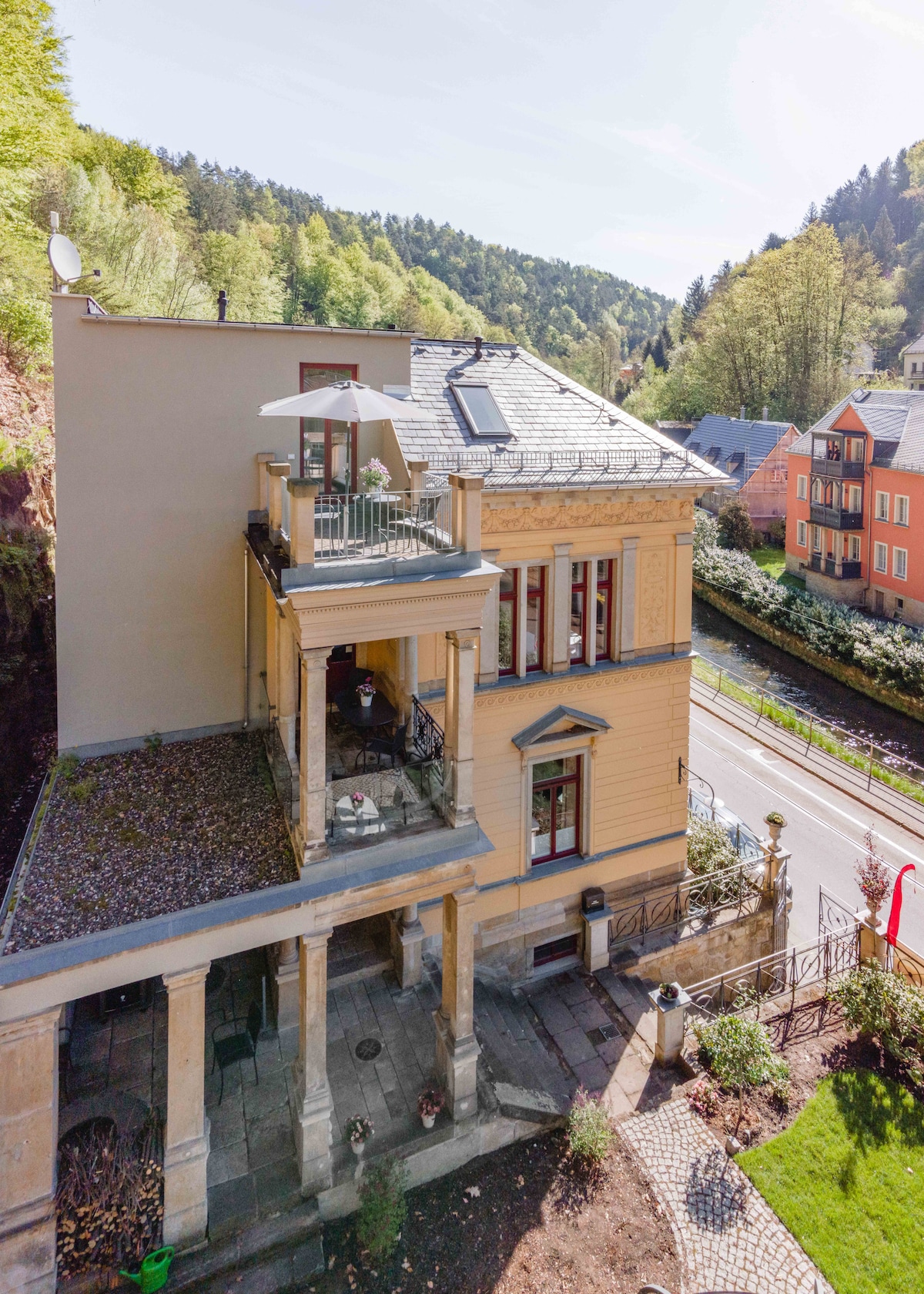 The exterior of Villa Emma is showcased, featuring classic architectural elements and a spacious balcony. Sunlight illuminates the building, highlighting its decorative details. Surrounding greenery offers a tranquil backdrop, while the nearby road is visible, connecting to the charming scenery of Bad Schandau.