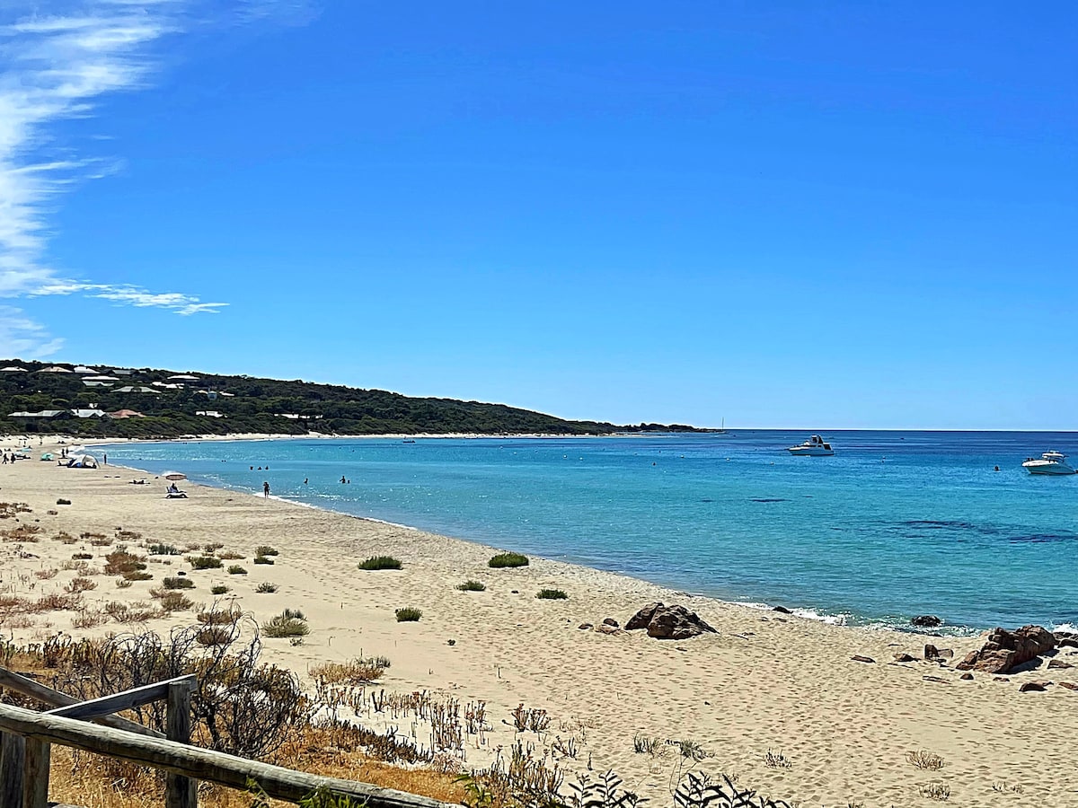 A serene beach scene captures the soft, golden sand meeting clear, turquoise waters. Gentle waves lap against the shore with a few people enjoying the sun. Lush greenery and distant coastal hills create a peaceful backdrop against a bright blue sky.