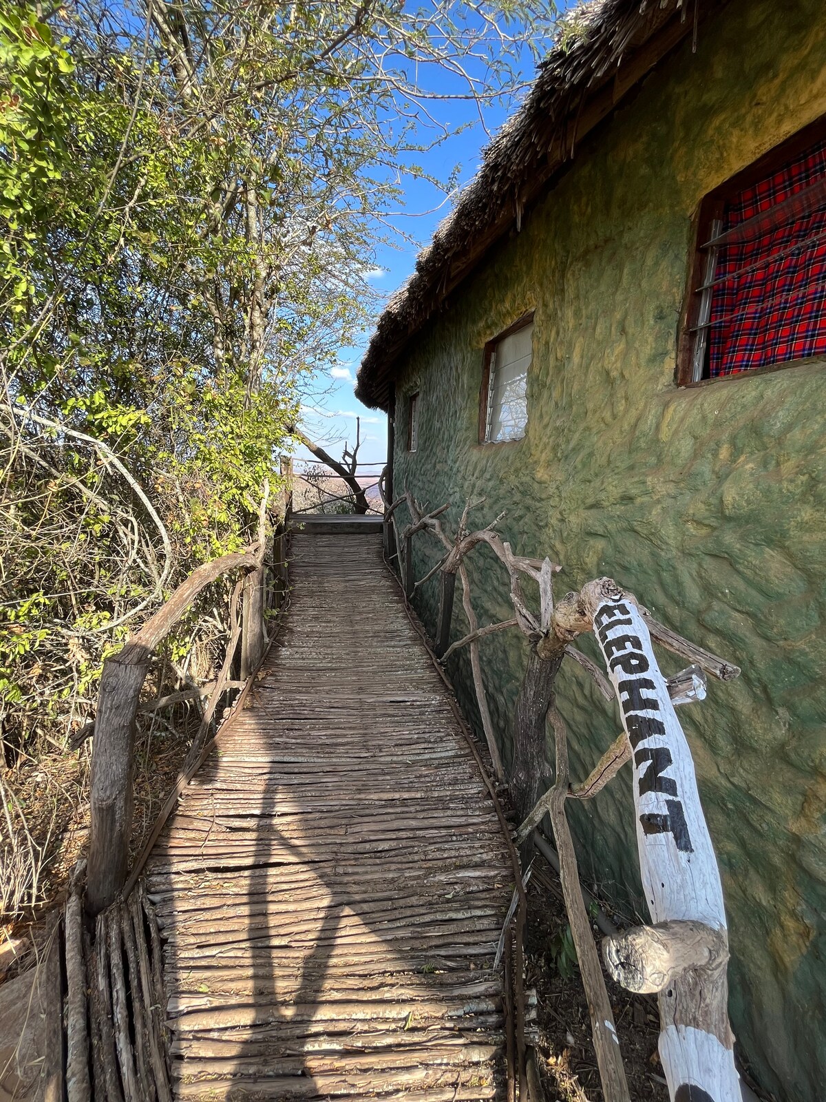 A wooden pathway, surrounded by lush greenery, leads to a textured, earth-toned hut with a thatched roof. Natural elements are incorporated into the design, highlighted by a hand-painted sign labeled 'ELEPHANT' affixed to the side of the walkway.