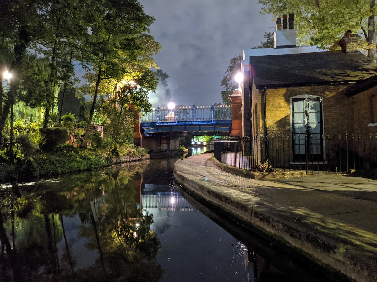 The image depicts a serene canal scene illuminated by soft lights. Reflections of trees and the nearby bridge are visible in the calm water. The pathway beside the canal features a stone surface, bordered by a decorative black wrought iron fence, creating an inviting atmosphere.