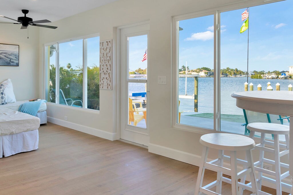 A bright and airy indoor space features large windows showcasing scenic water views. A seating area with a daybed and a compact dining space is visible, complemented by a bar with stylish stools. The doors open to a waterfront patio, inviting natural light.