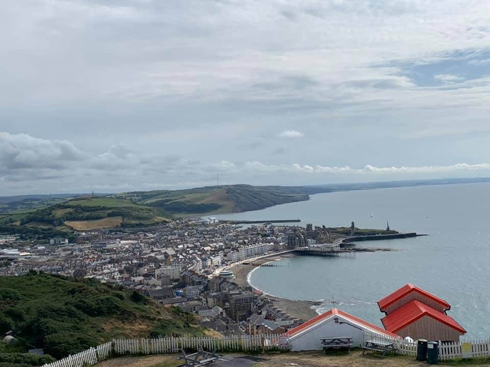 A panoramic view captures the coastline with the town nestled along the beach. The sea curves gracefully around the shore, while the distant hills provide a lush backdrop. Two red-roofed structures are positioned in the foreground, overlooking the scenic landscape.