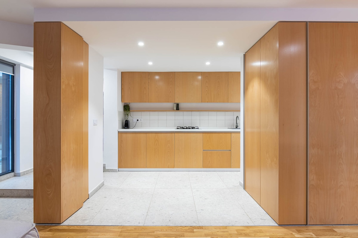 A modern kitchen area is enclosed by sleek wooden cabinetry, featuring white countertops and minimalist shelf space. Bright lighting highlights the open layout. A stovetop is visible, and natural light is welcomed through large glass doors, connecting to the surrounding environment.