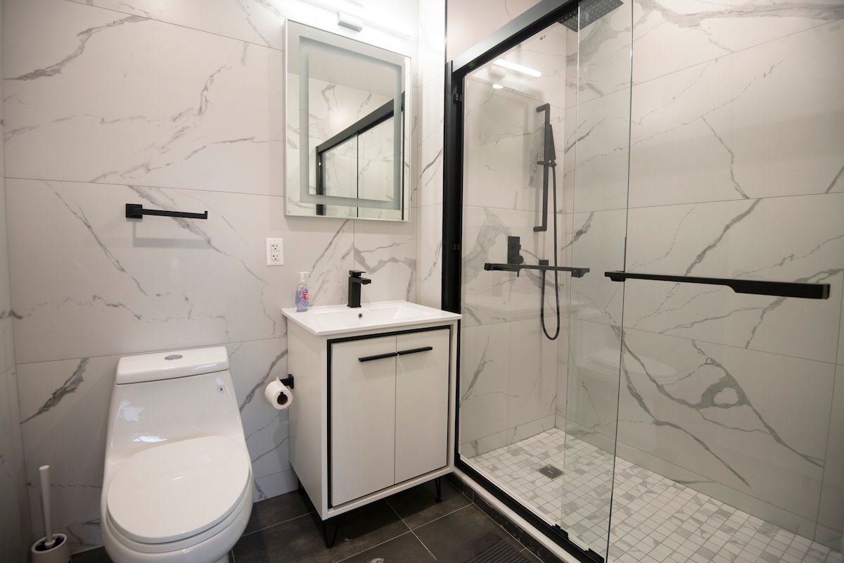 A modern bathroom features elegant marble tiles with a spacious glass shower enclosure. A contemporary vanity with a mirror is positioned beside a toilet. Minimalistic black fixtures add a sleek touch to the clean design, enhancing the overall functionality of the space.