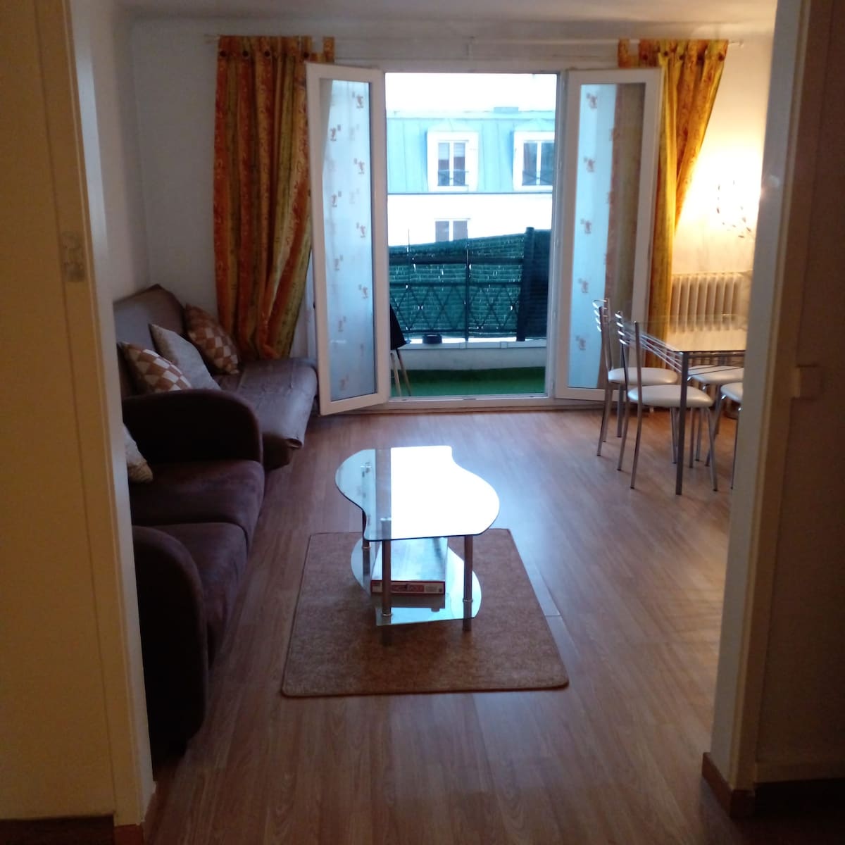 An open living area features a brown sofa accompanied by decorative cushions. A glass coffee table is positioned on a light area rug. Dark curtains frame large double doors that lead to a small balcony, while a dining table with chairs is visible in the background.