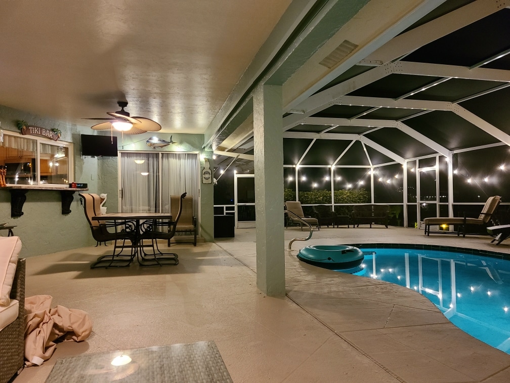The pool area is enclosed by a screened lanai, offering a relaxing space with comfortable seating. A dining table and chairs are positioned nearby, alongside lounge chairs. Soft lighting enhances the ambiance around the heated pool, creating an inviting environment for gatherings.