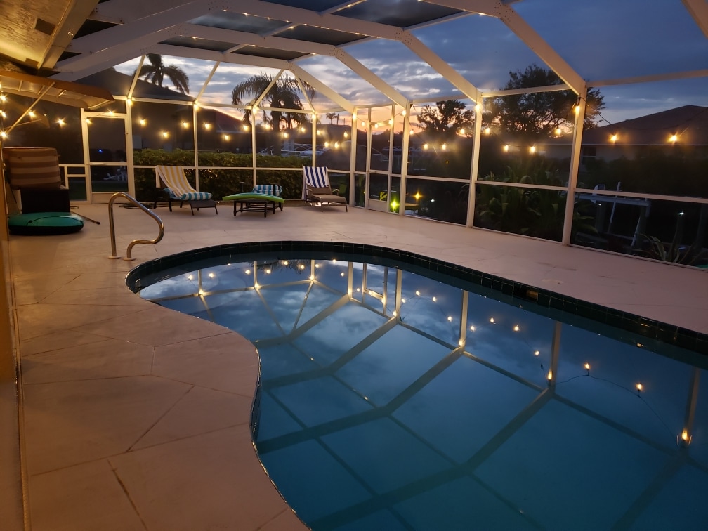 A serene pool area features a heated pool surrounded by a spacious deck. Soft lighting enhances the atmosphere, while lounge chairs are positioned for relaxation. A protective screen enclosure is visible, and lush greenery frames the area, providing a tranquil outdoor retreat.