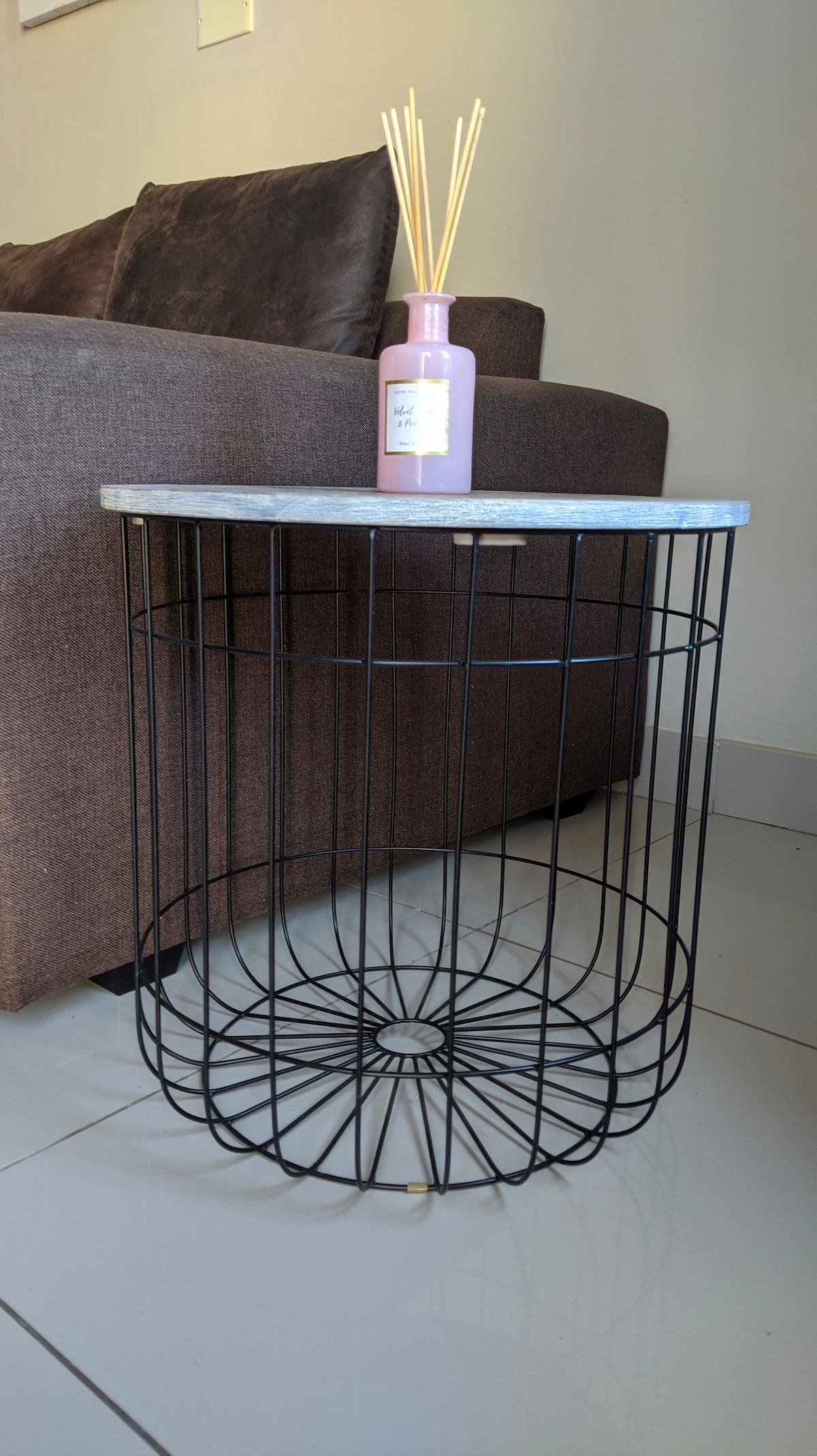 A stylish side table featuring a round marble top supported by a black wire frame. A decorative bottle, tinted pink, is placed on the table alongside a set of reed diffuser sticks. The backdrop includes a brown sofa, enhancing the modern design.