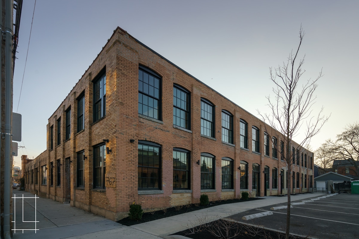 The exterior of the loft building is showcased, featuring red brick walls and large, modern windows. The structure has a distinct industrial design with multiple defined sections and a pathway leading to the entrance. A small tree is visible in the foreground.
