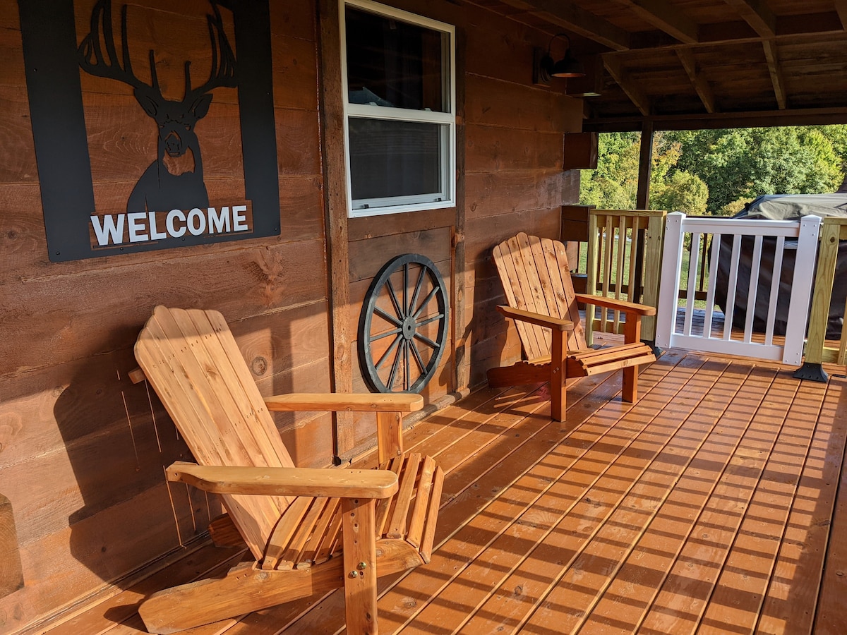 Two wooden Adirondack chairs are positioned on the covered porch, inviting relaxation. A rustic 'WELCOME' sign and a decorative wheel add charm to the warm wooden facade. Natural light filters through the window, enhancing the cozy outdoor space.