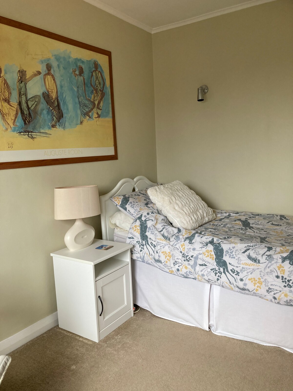 A cozy bedroom features a comfortable single bed, dressed in floral bedding and topped with a textured cushion. A bedside table with a lamp is positioned alongside the bed, and a colorful artwork adorns the wall, adding character to the neutral-toned room.