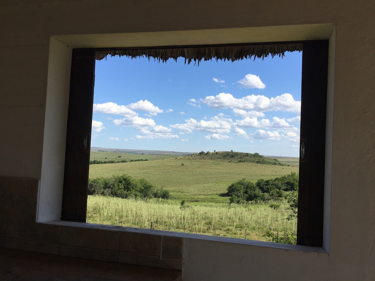 A large window frames a sweeping view of the surrounding countryside, filled with lush green fields and rolling hills. White clouds drift across a bright blue sky, providing a serene backdrop to the tranquil landscape.