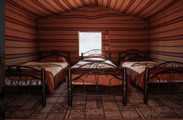 A spacious tent interior is depicted with four beds, each featuring dark metal frames and warm-toned bedding. The walls are adorned with colorful striped fabric, and a single window allows natural light to enter, illuminating the patterned floor.