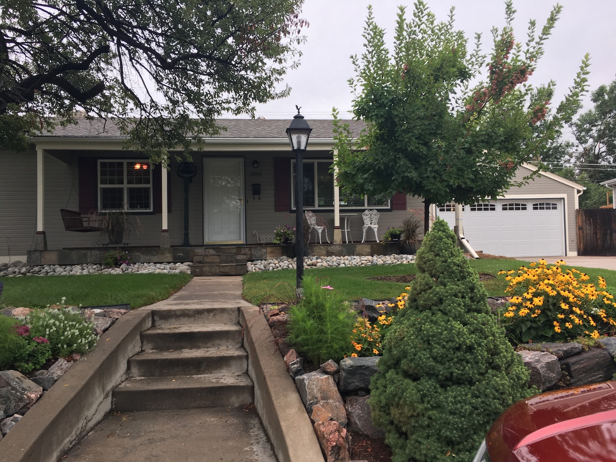 A welcoming front yard features a stone pathway leading to the entrance of the house. Lush greenery and colorful flowers are arranged around the yard. The porch is furnished with white chairs, and a lamp post enhances the inviting appearance of the exterior.