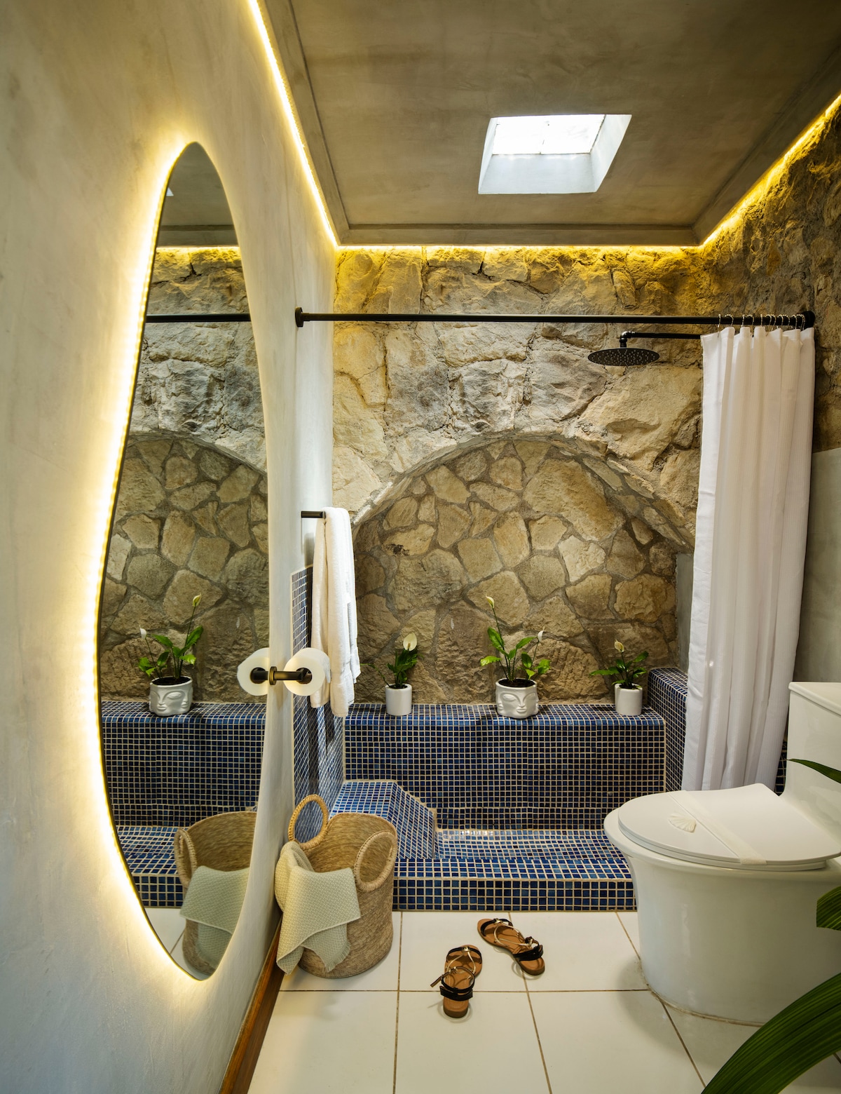 The bathroom showcases an organic-shaped full-length mirror illuminated by indirect lighting. A stone wall feature complements the serene atmosphere. A shower with a rain showerhead is visible, alongside a modern toilet. Two potted plants add a touch of greenery to the space.