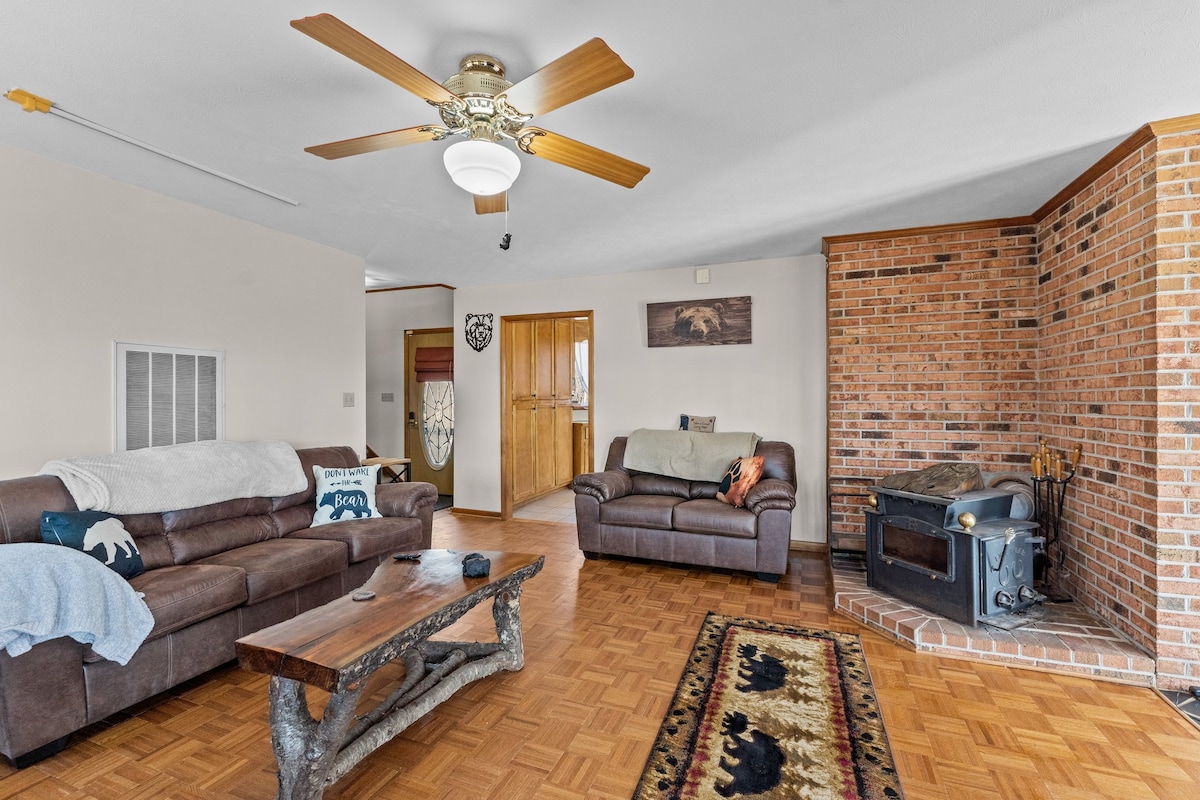 A welcoming living area features two leather couches and a rustic wooden coffee table. A charming stone fireplace with a wood stove adds warmth, while a ceiling fan provides airflow. Natural light fills the space through nearby openings, hinting at the home's cozy atmosphere.