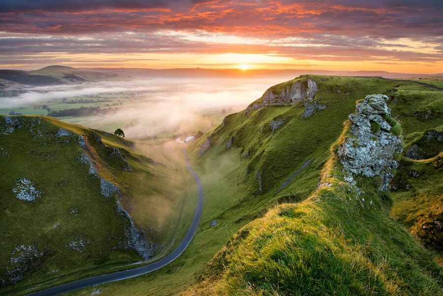 A scenic view features rolling hills and a winding road cutting through lush green valleys. Soft morning mist lingers, while the sky is painted with warm colors of sunrise. The landscape showcases a tranquil and open countryside, inviting exploration of the natural beauty.