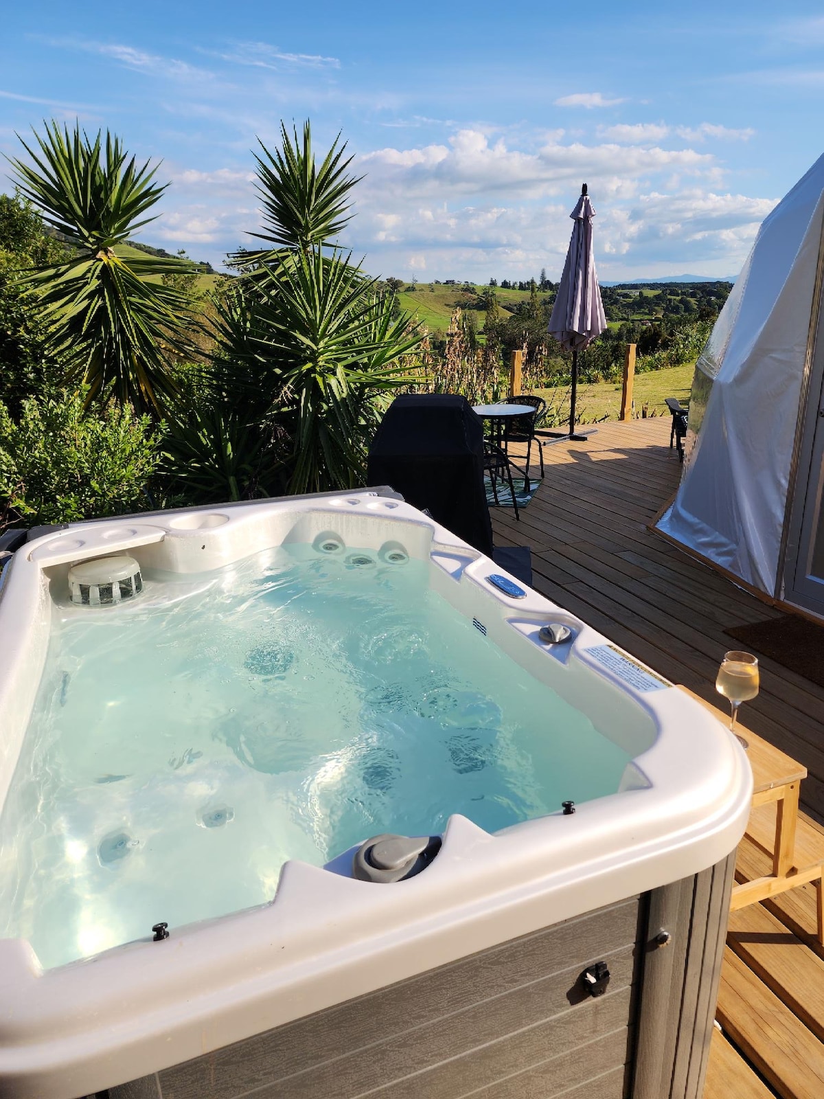 A private hot tub sits at the edge of a wooden deck, offering views of the surrounding greenery and hills. A glass of wine rests on a nearby wooden stool, and a shaded patio umbrella stands in the distance, adding to the outdoor relaxation setting.