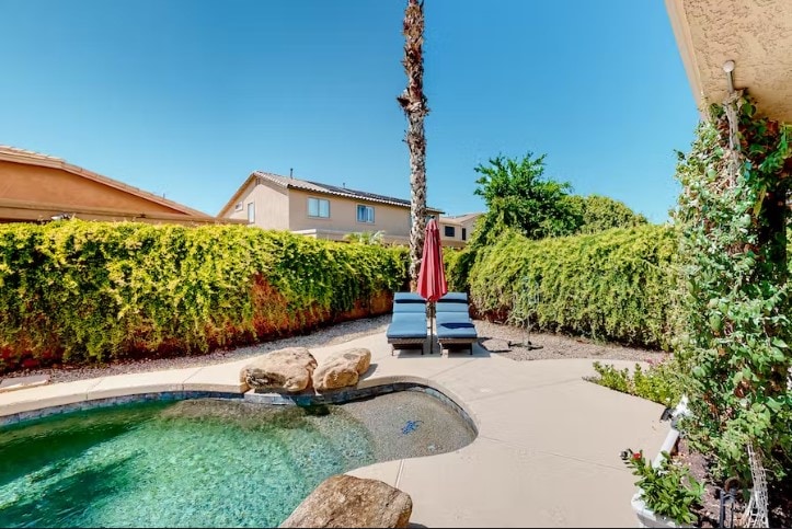 A private backyard hosts a saltwater pool surrounded by lush greenery. Two lounge chairs with red umbrellas provide shaded seating, while a rock feature enhances the natural landscape. The clear blue sky above complements the tranquil outdoor space.