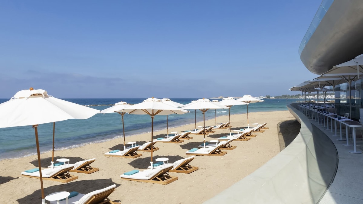A sandy beach is lined with several sun loungers under white umbrellas. The gentle waves of the sea can be seen in the background, stretching towards a clear blue sky. A curving modern building is partially visible at the edge of the beach.