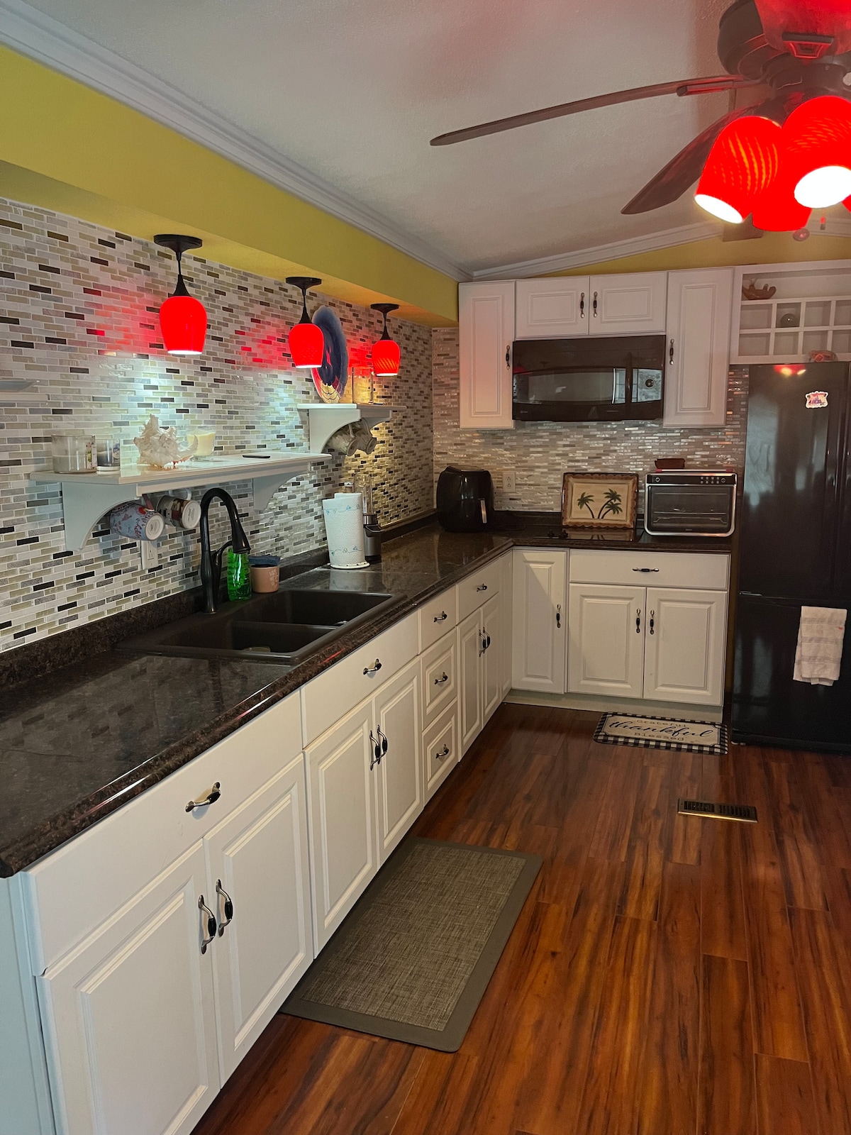 A well-equipped kitchen is presented with dark granite countertops and white cabinetry. Red pendant lights create a distinctive glow over the workspace. Modern appliances include a black refrigerator and a microwave, while a textured backsplash adds visual interest.