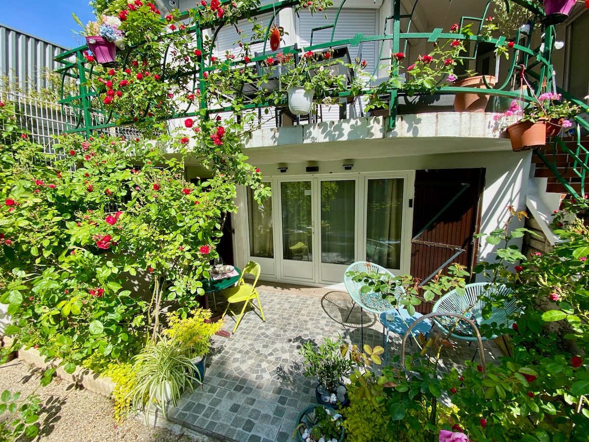 The outdoor space is surrounded by lush greenery and vibrant flowers, featuring a cozy patio with colorful chairs and a small table. The entrance to the studio is framed by plants, creating a serene and welcoming atmosphere.